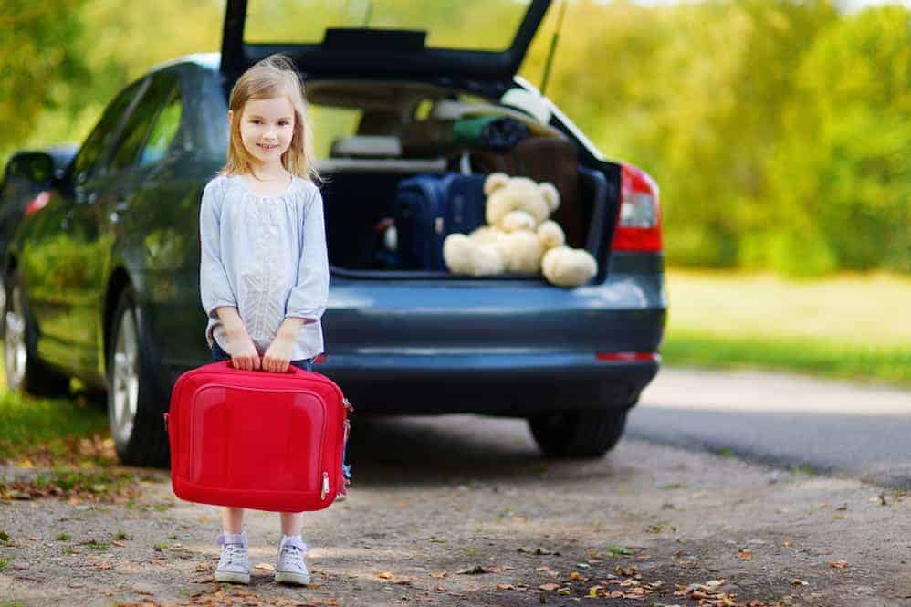 Little girl with a red suitcase standing in front of a car.